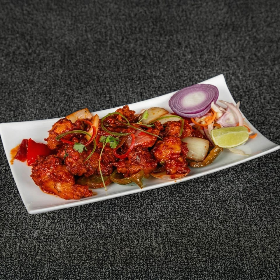 Spicy red chicken dish with bell peppers, onions, cilantro, and a lime wedge on a white plate.