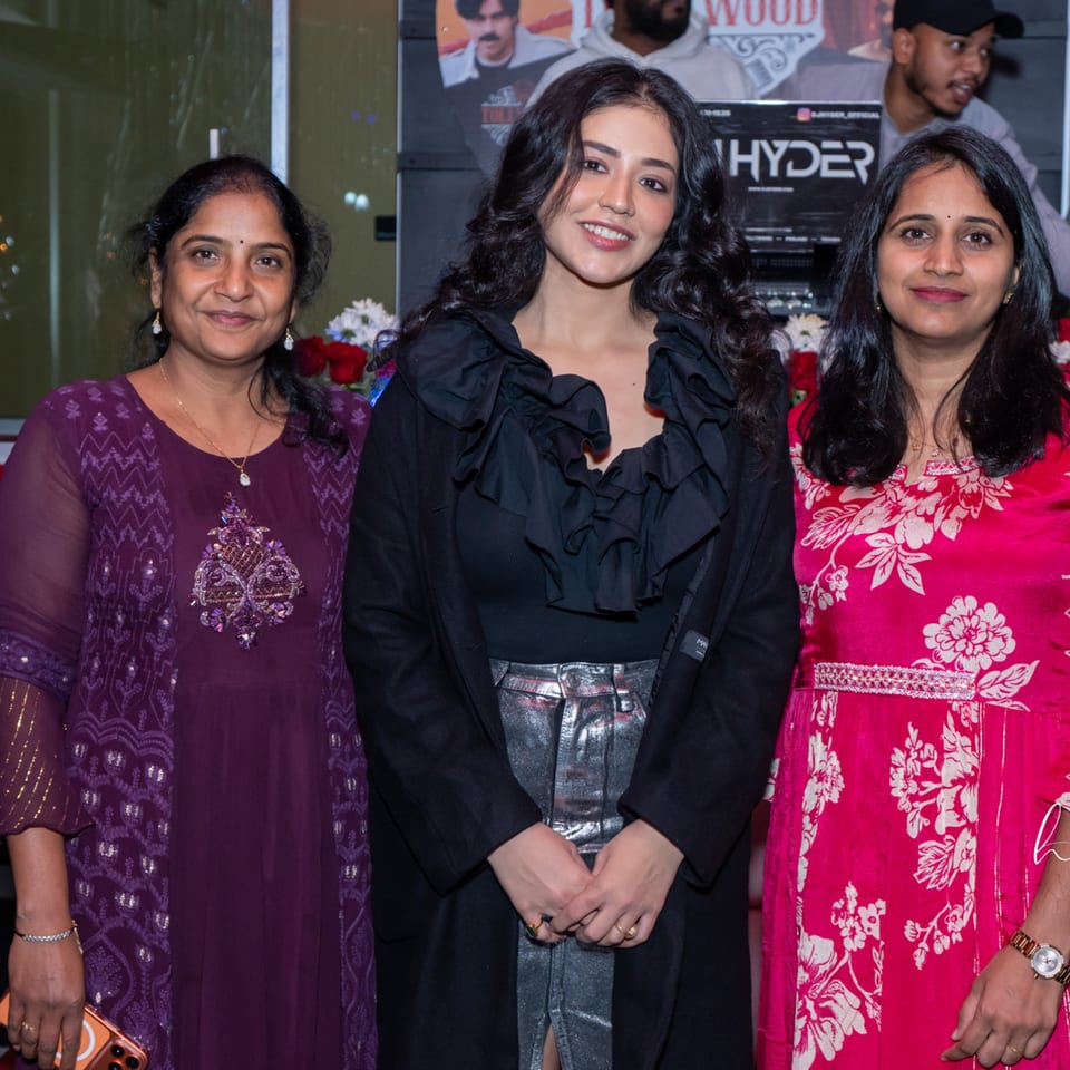 Three women smiling at the camera, with a man and a "HYDER" sign in the background.