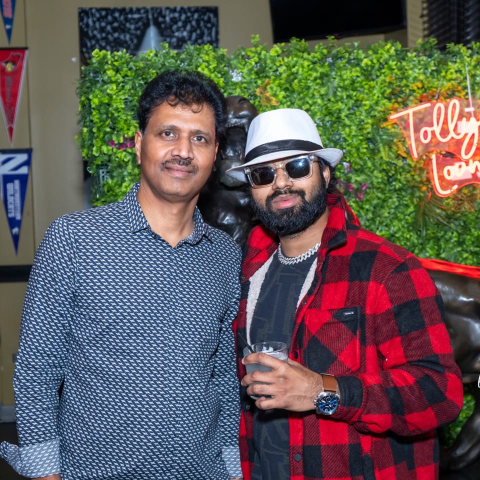 Two men posing for a photo in front of a green wall with a neon sign.