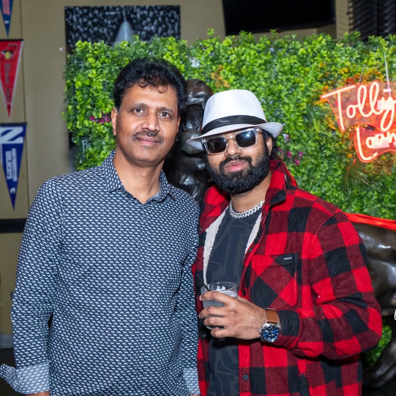 Two men posing for a photo in front of a green wall with a neon sign.
