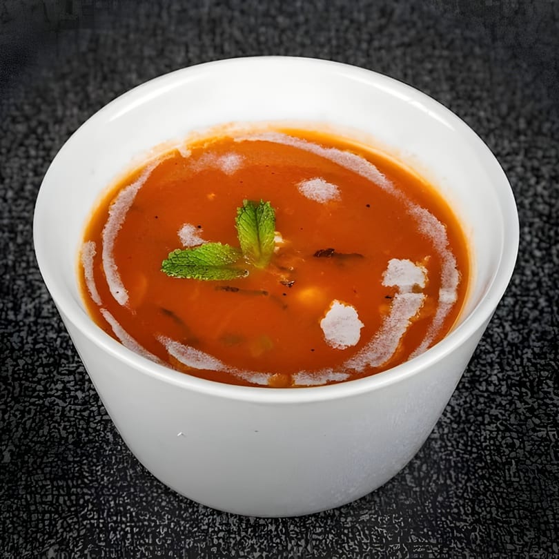A white bowl of tomato soup with cream swirls and a mint garnish on a dark textured surface.