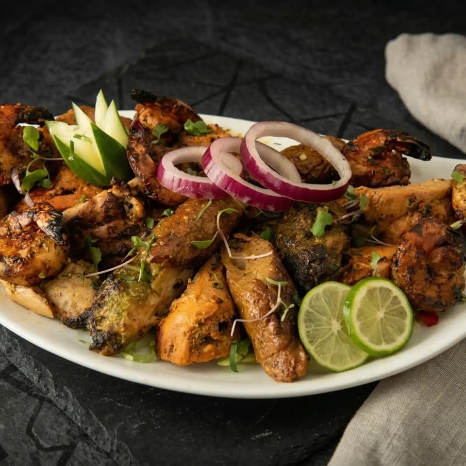 Grilled chicken, shrimp, and sausage with red onion, lime, and cucumber on a white platter.