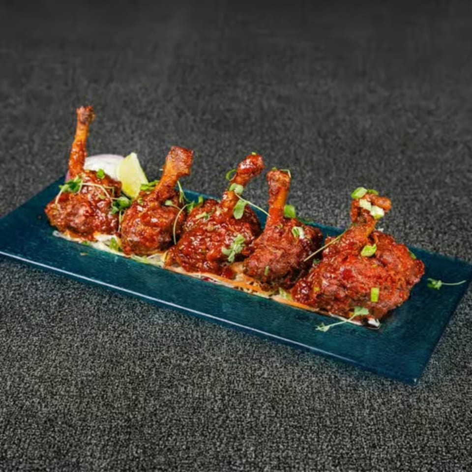 Five chicken lollipops with red sauce, garnished with herbs and lime, on a blue rectangular plate.