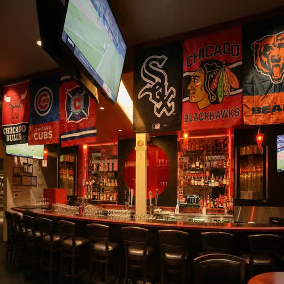 A sports bar with Chicago team flags, a long bar with stools, and TVs showing a game.